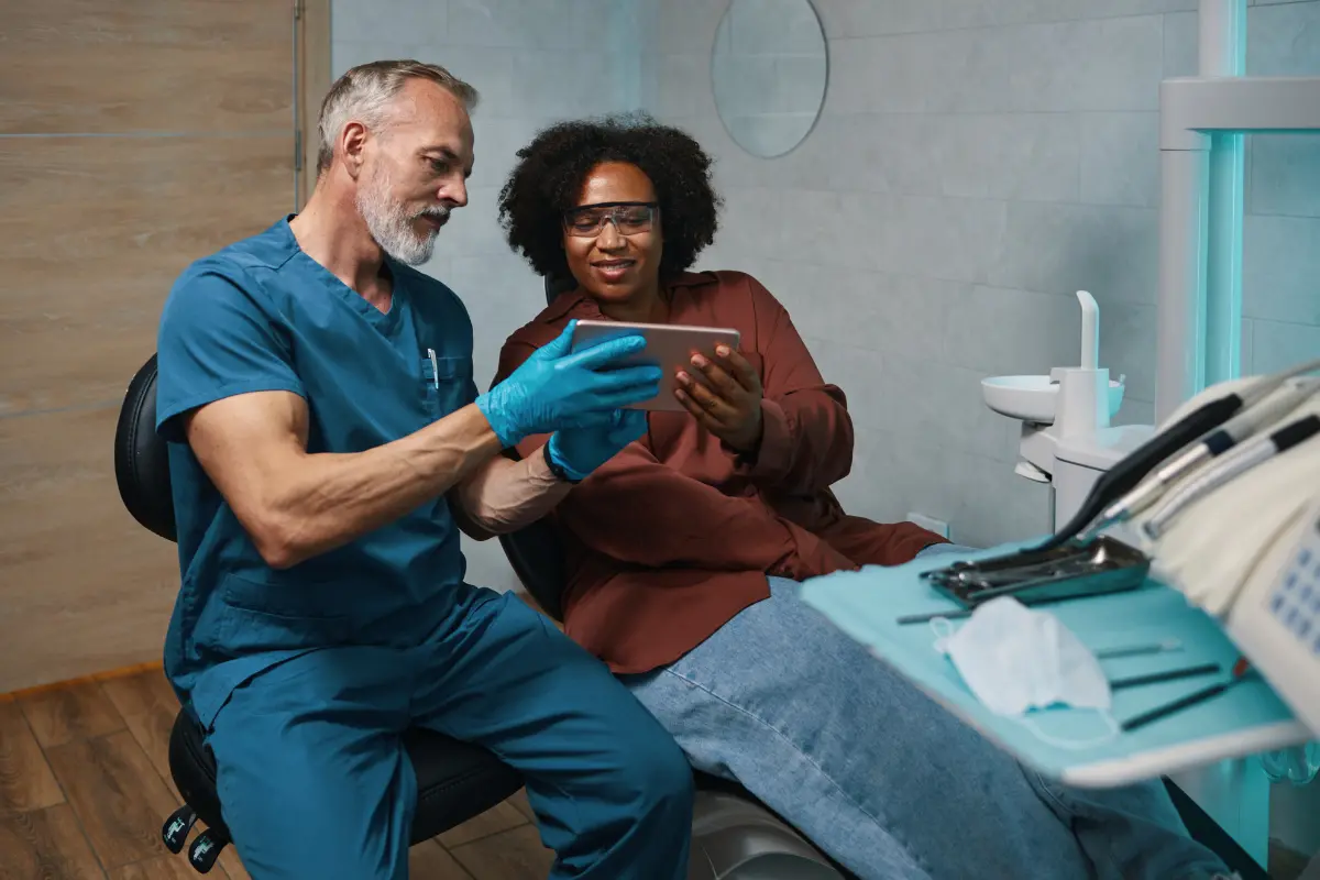 Zahnarzt in blauer Arbeitskleidung und blauen Handschuhen zeigt einer Patientin mit Schutzbrille etwas auf einem Tablet in einer modernen Zahnarztpraxis.