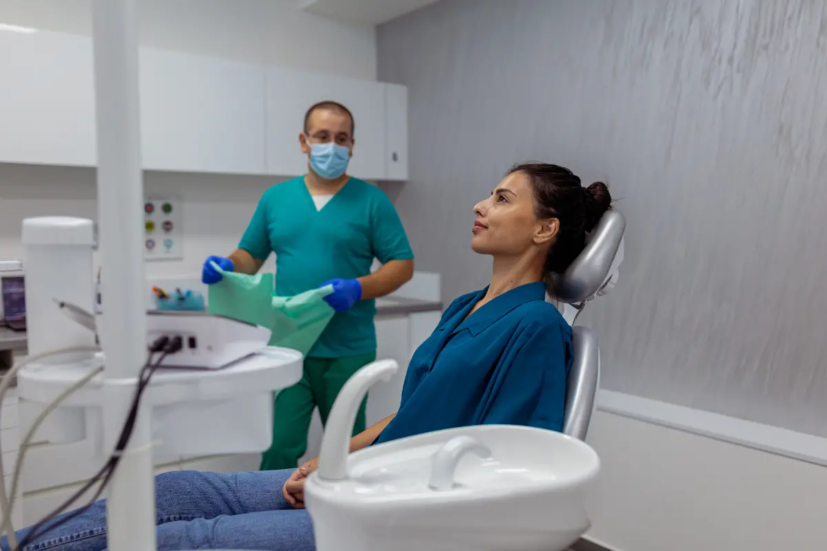 Zahnarzt in grüner Berufskleidung und Maske bereitet den Behandlungsraum vor, Patientin sitzt entspannt im Behandlungsstuhl