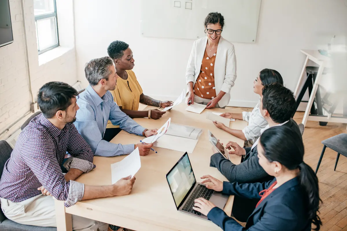 Diverse Gruppe von sechs Personen bei einem Meeting an einem Tisch, eine Frau steht präsentierend mit Unterlagen in der Hand