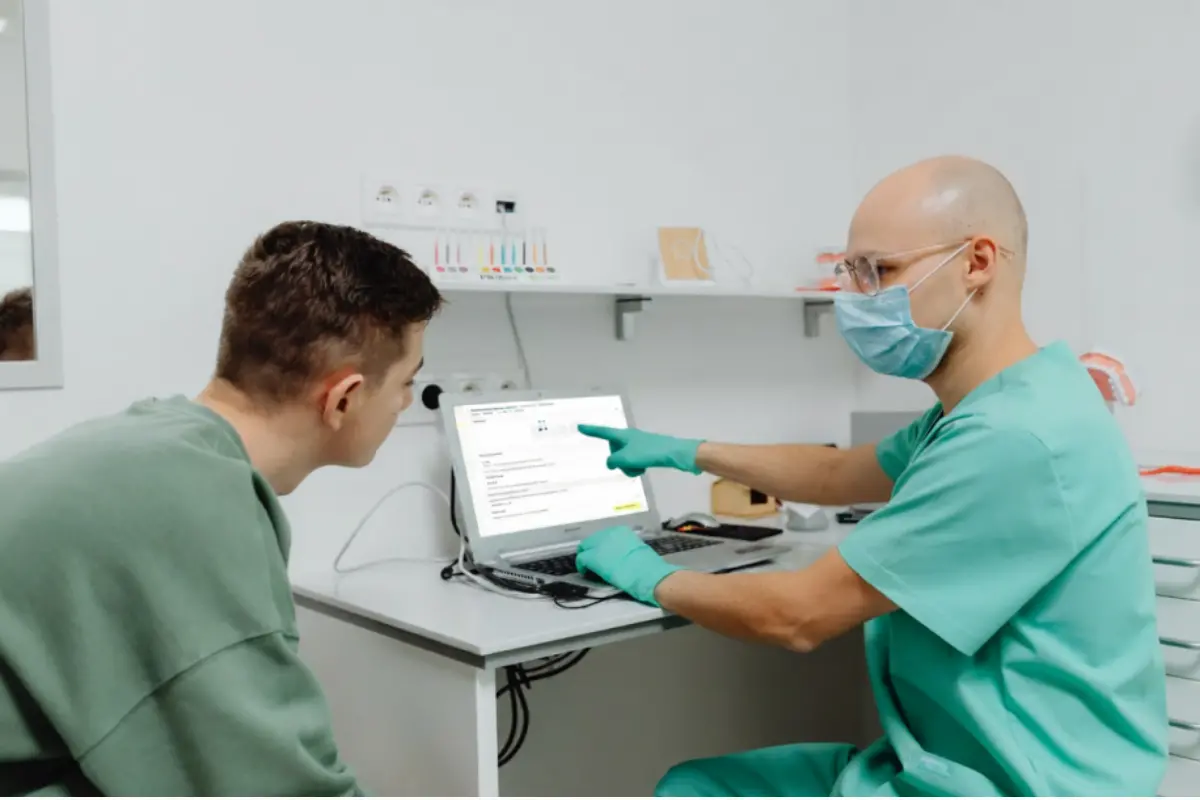 Zahnarzt mit Maske und grünen Handschuhen erklärt einem Patienten einen Behandlungsplan auf einem Laptop-Bildschirm in der Zahnarztpraxis.