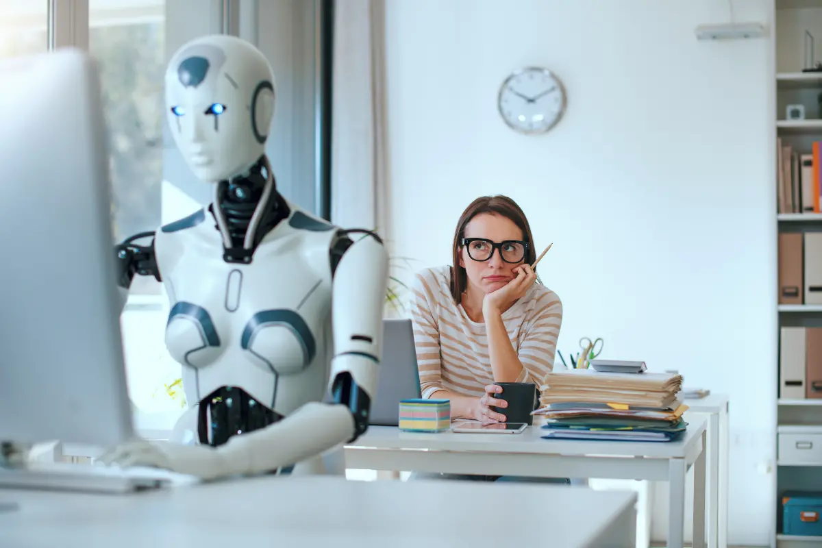 Frau mit Brille sitzt nachdenklich an einem Schreibtisch neben einem humanoiden Roboter vor einem Computer