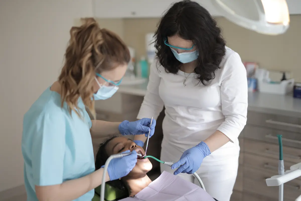 Two dental professionals treating a female patient in a dental chair, both wearing face masks and blue disposable gloves, in a modern dental practice.