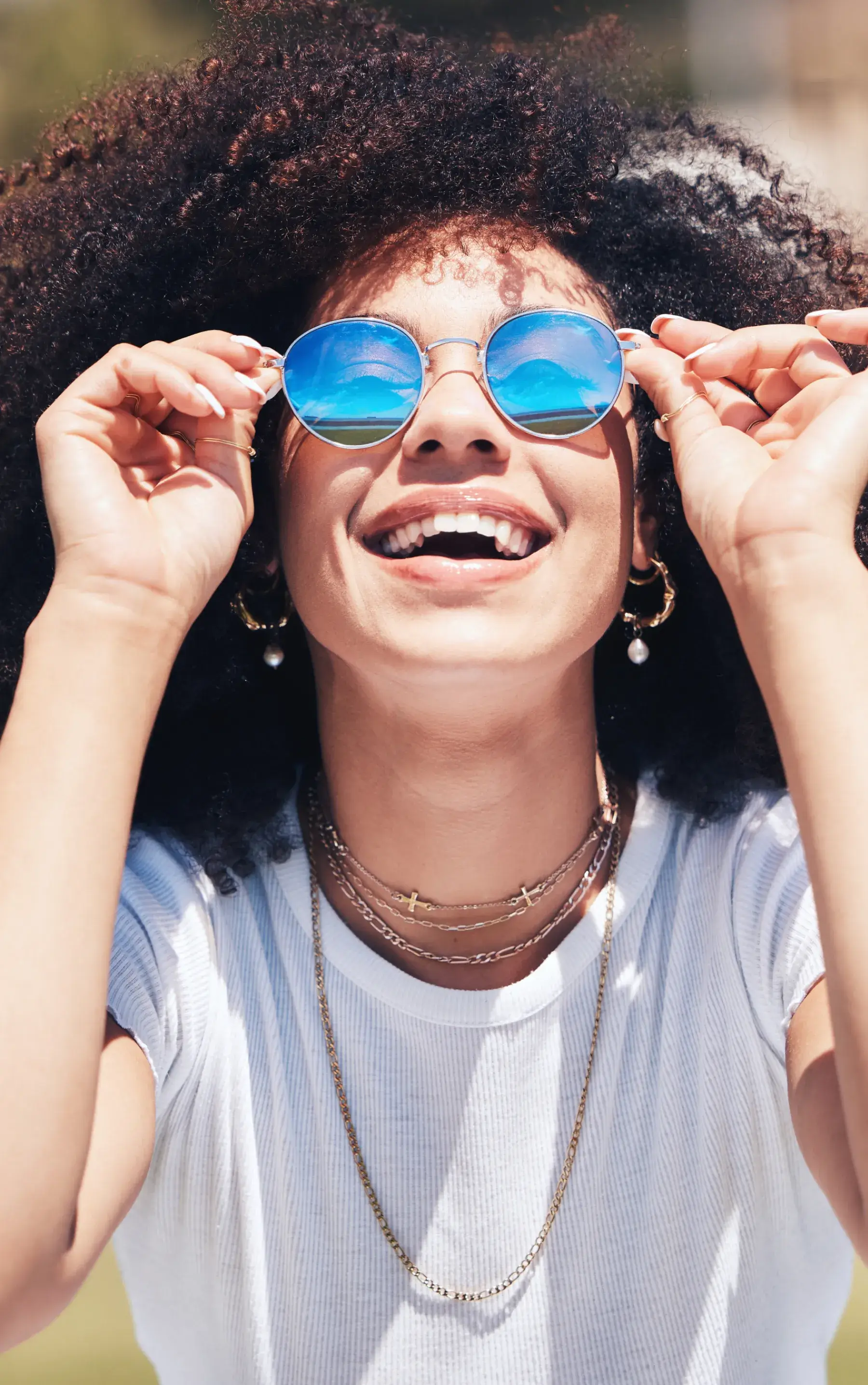 Smiling woman adjusting blue reflective sunglasses with curly hair and wearing layered gold necklaces and hoop earrings.