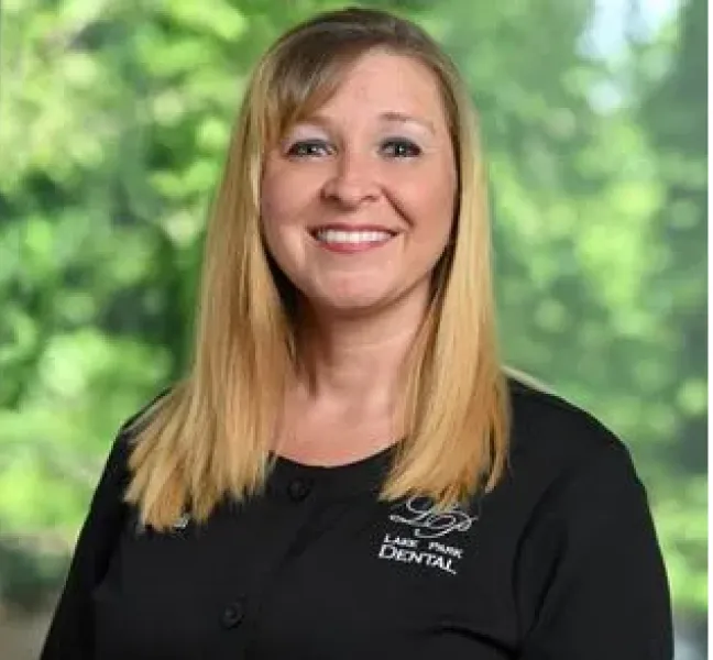 Smiling woman with straight blonde hair wearing a black dental uniform with Lake Park Dental logo against a green blurred background.