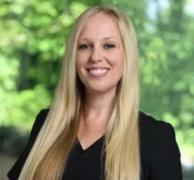 Smiling woman with long blonde hair wearing a black top, standing outdoors with green foliage in the background.
