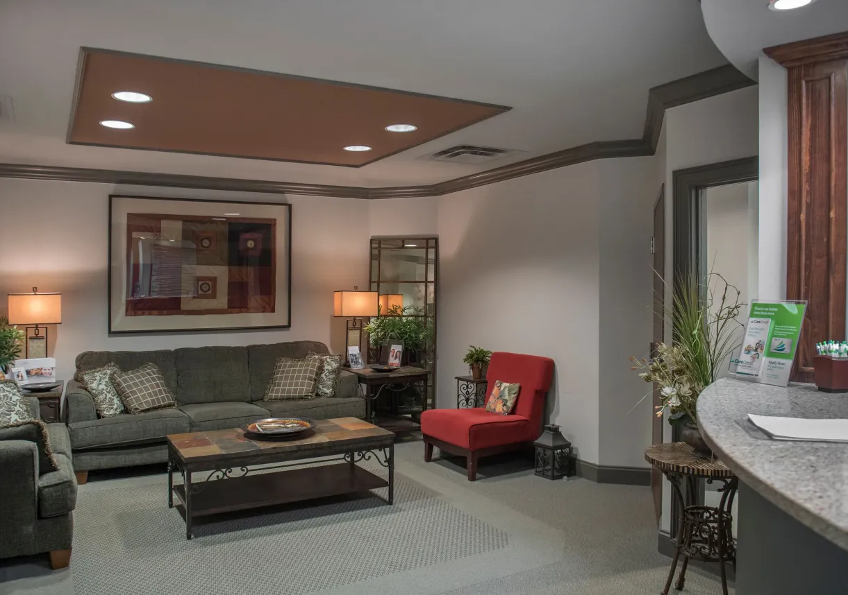 Cozy office waiting area with gray sofas, a red chair, a coffee table, lamps, and decorative plants.