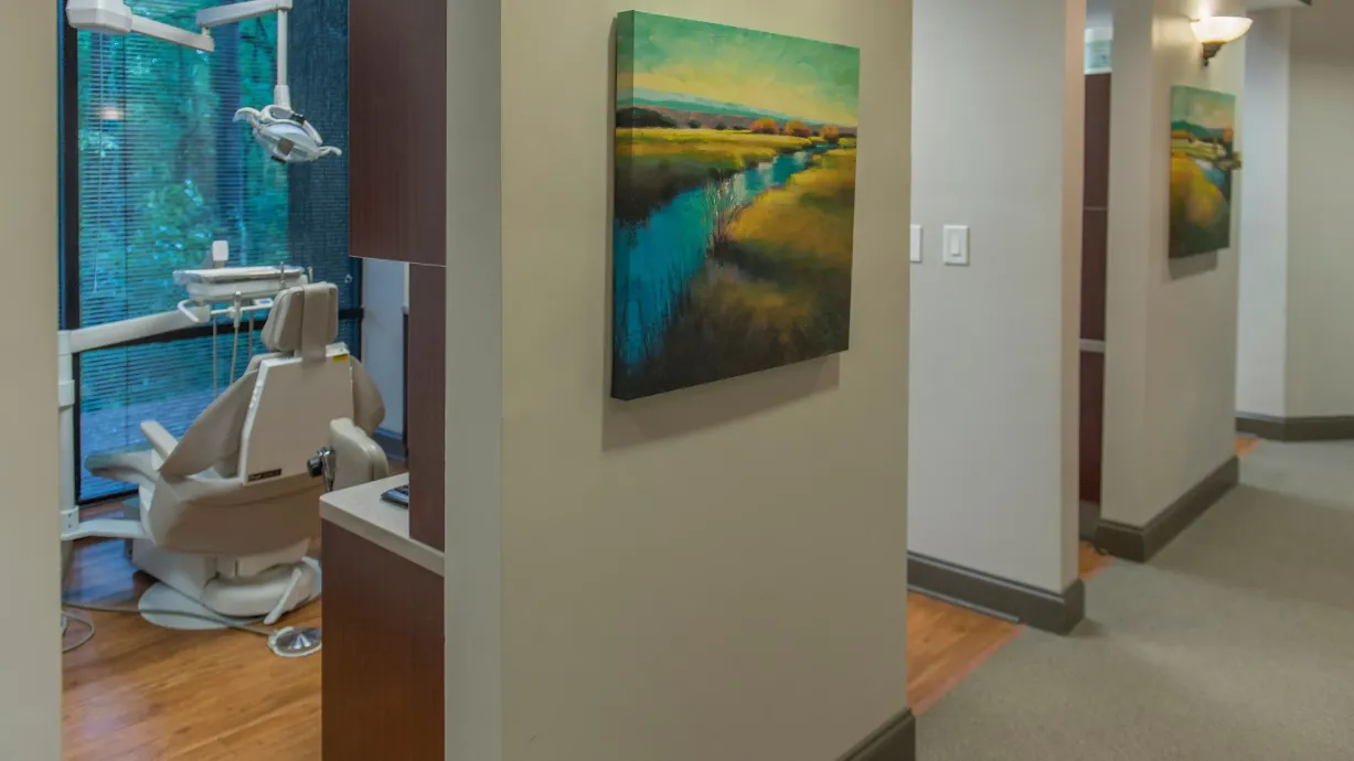 Hallway in a dental office with beige walls, a landscape painting, and a dental chair visible through an open door.