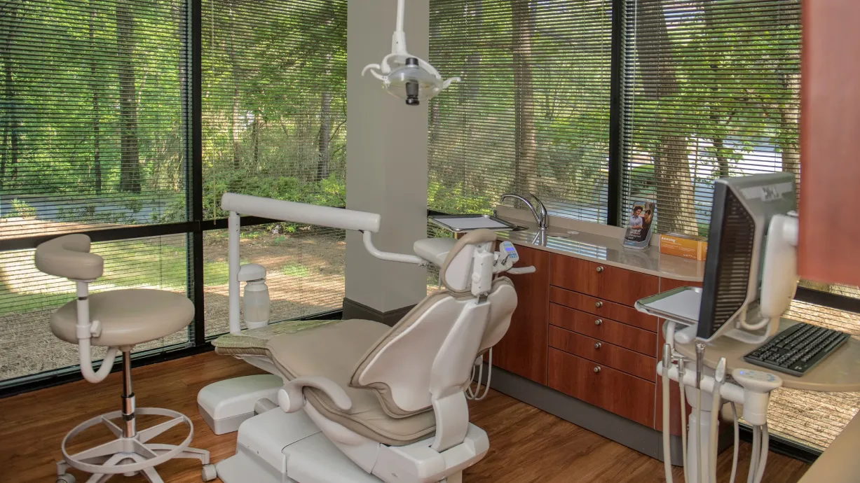 Modern dental office with beige dental chair, stool, countertop with sink, and computer, surrounded by large windows with blinds showing trees outside.