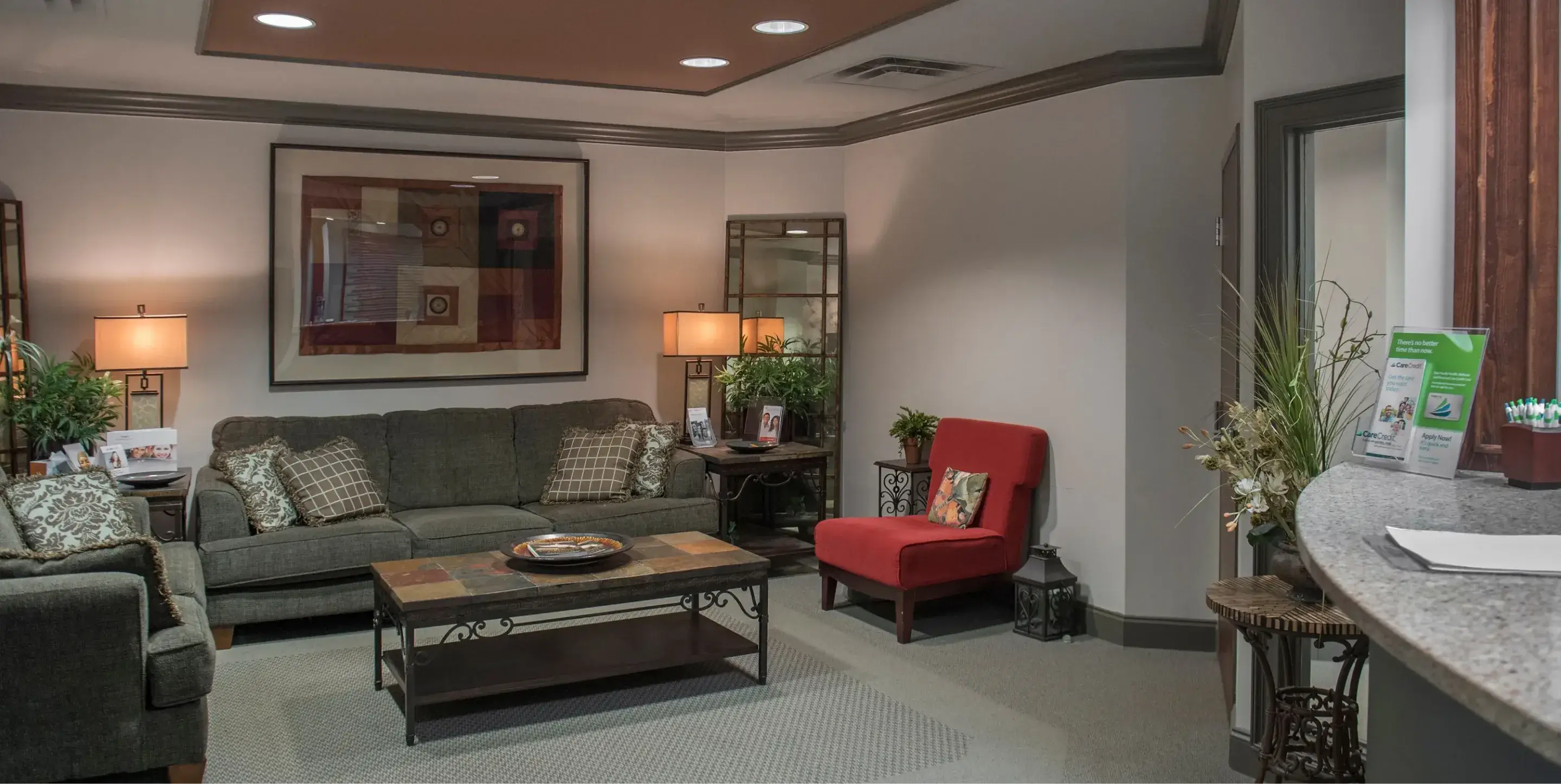 Cozy office lounge with grey sofas, a red armchair, coffee table, lamps, decorative plants, and framed artwork on the wall.