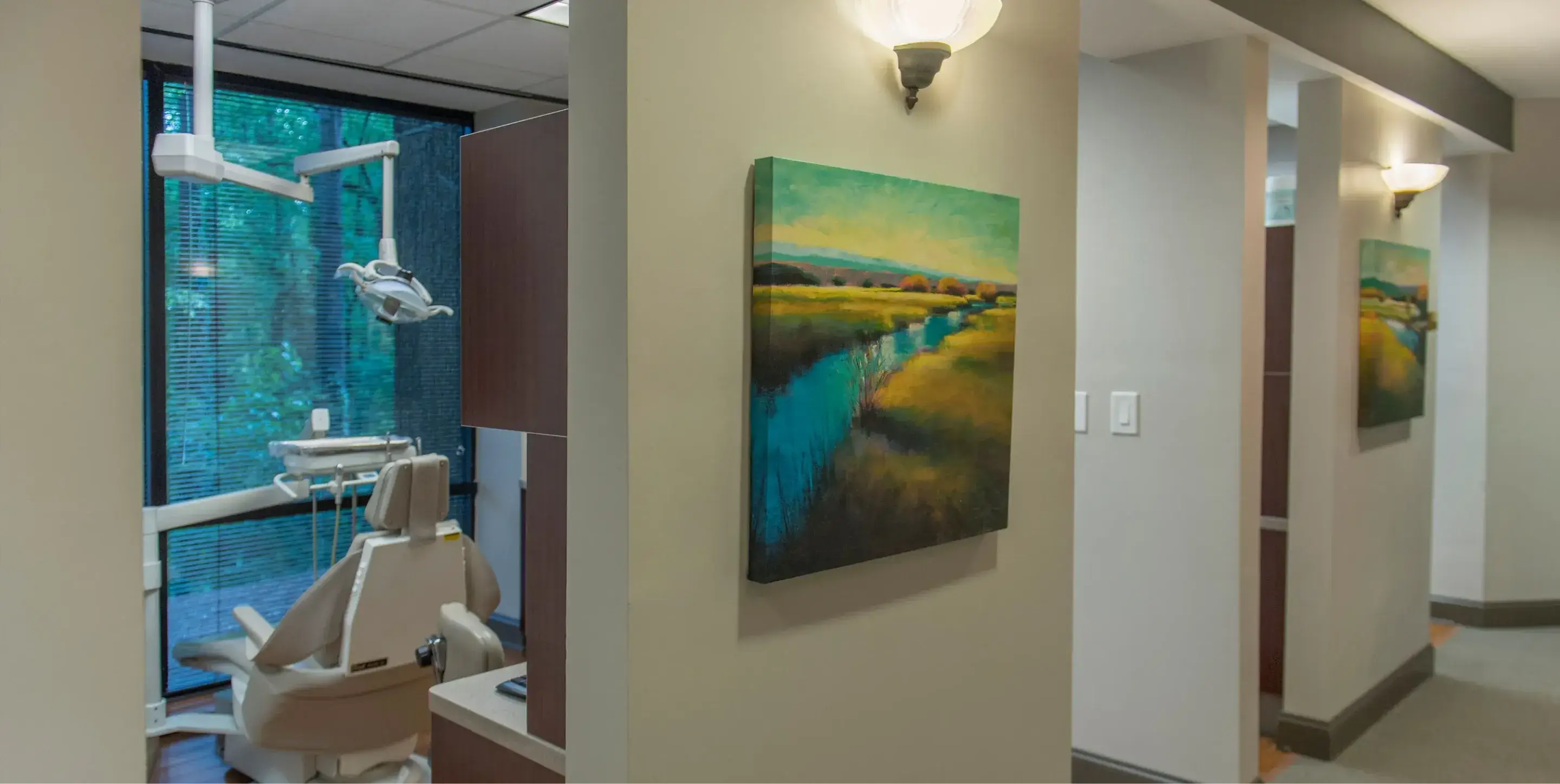 Dental chair and equipment next to a hallway wall with a landscape painting and wall-mounted light above it.