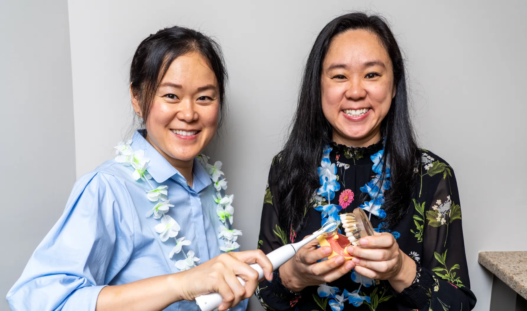 Two smiling women wearing floral leis demonstrating an electric toothbrush on a dental model.
