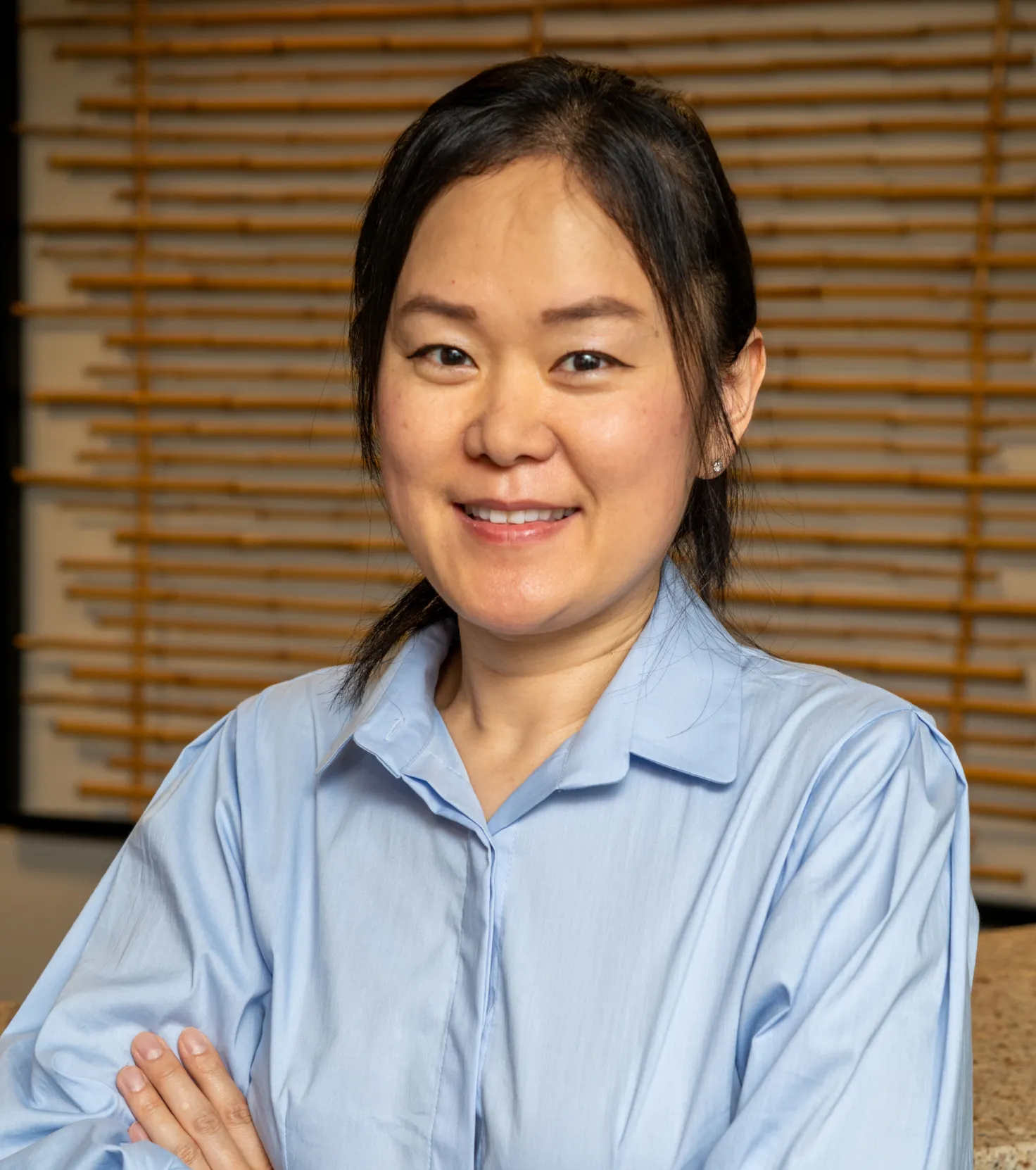 Smiling woman with dark hair wearing a light blue button-up shirt in front of a wooden slatted background.