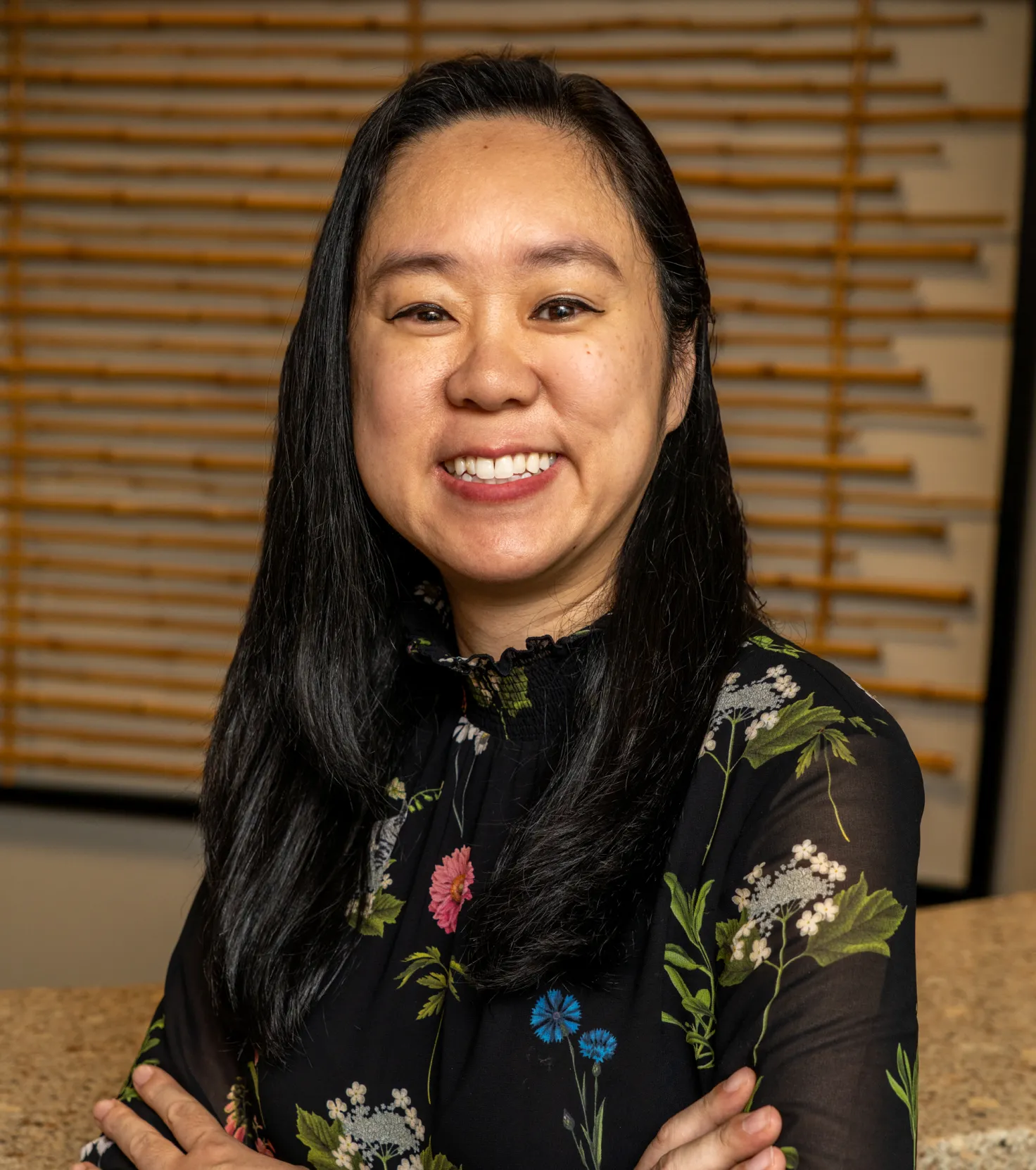 Smiling woman with long black hair wearing a black floral blouse, standing with arms crossed.