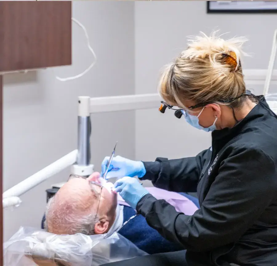 Dentist wearing a mask and gloves examining an elderly male patient's teeth in a dental clinic.