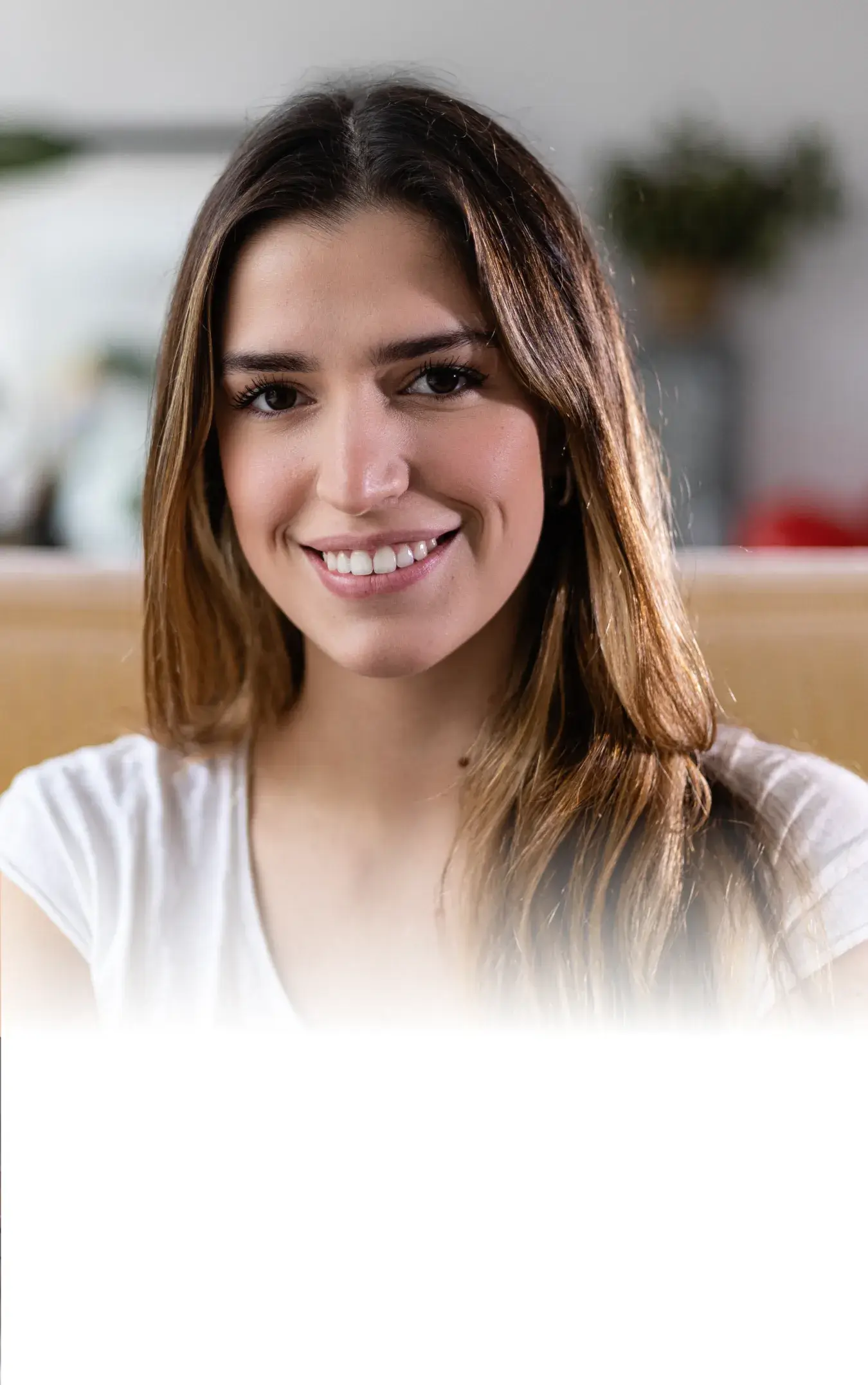 Young woman with long brown hair smiling, wearing a white shirt, with blurred indoor background.