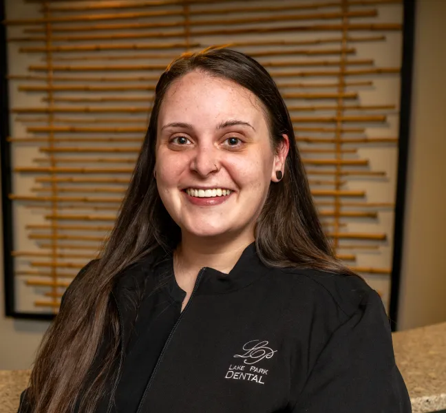 Smiling woman with long brown hair wearing a black Lake Park Dental jacket in front of a wooden decorative wall.