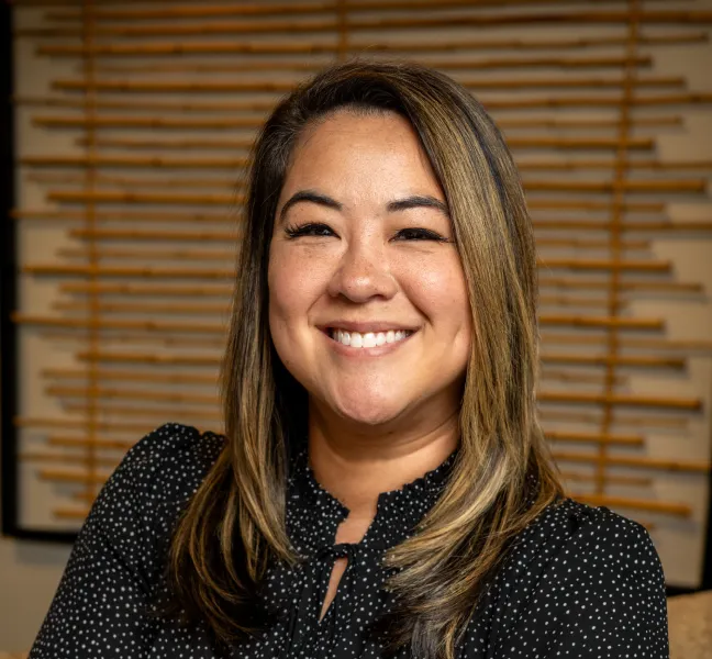 Smiling woman with shoulder-length light brown hair wearing a black blouse with white polka dots, posed against a wooden slat background.