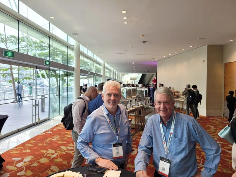 Mark Lay and Al Giebel, VP of Marketing at Arcadian, smile for the camera during the ATS Conference 2024 in Singapore. The photo is taken in a bright convention hall with floor-to-ceiling windows and a buffet setup in the background.