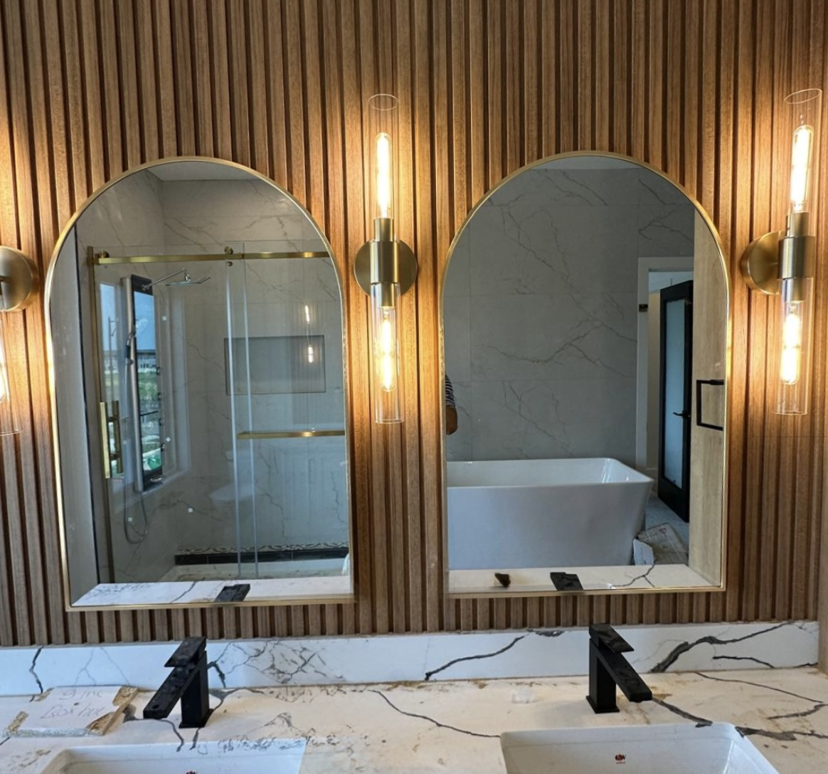 Two arched brass-framed custom mirrors installed above a marble double vanity in a modern Edmonton bathroom, flanked by gold tube wall sconces on a wood slat feature wall — custom mirror installation by OZ Shower Doors.