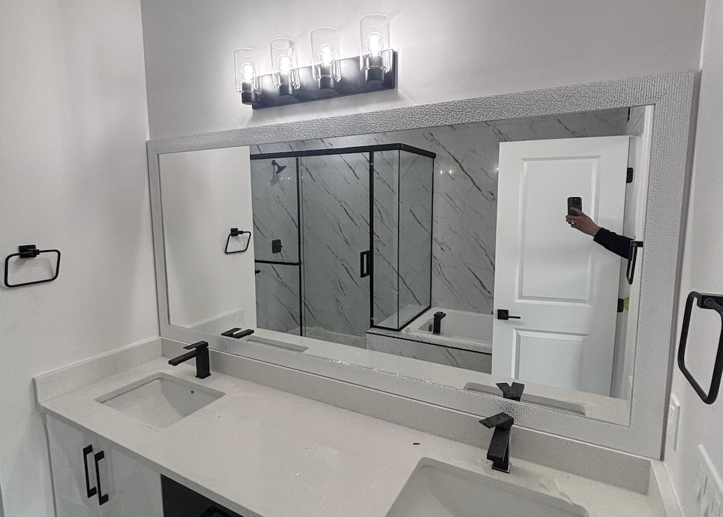 Large custom framed vanity mirror with a textured silver frame above a white quartz double-sink vanity in a modern Edmonton bathroom, reflecting a marble-tiled walk-in shower — custom mirror installation by OZ Shower Doors.