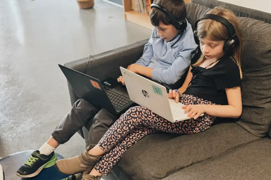 two students sitting on a couch wearing headphones and using laptops