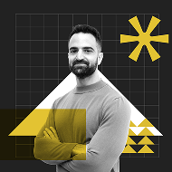 A man with a beard wearing a gray sweater stands confidently with arms crossed. The background features geometric shapes in yellow and a grid pattern.