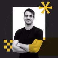 A man with a slight smile standing with arms crossed, wearing a black t-shirt. The background is graphic with geometric shapes in yellow and black, including a circular design and other patterns.