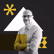 A man with glasses wearing a light gray shirt, arms crossed, smiling against a patterned black background with geometric shapes in white and yellow.