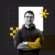 A man with glasses smiling, wearing a dark hoodie, poses with arms crossed against a black background with geometric yellow shapes and a white square.