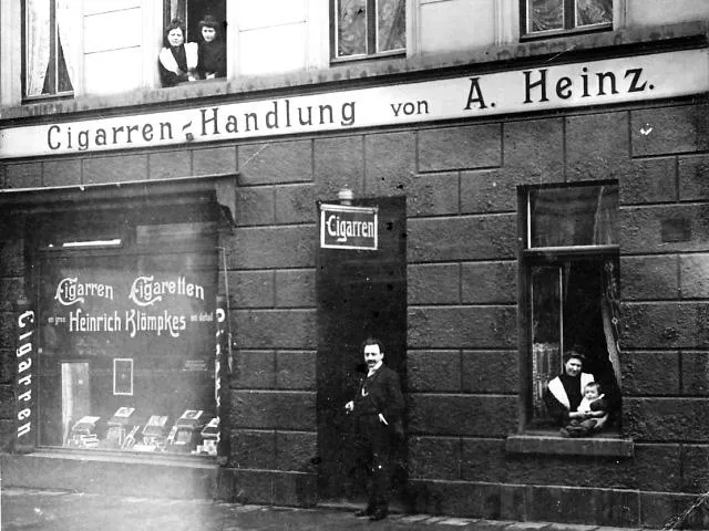 Schwarzweiß-Foto eines Tabakladens mit der Aufschrift ‚Cigarren-Handlung von A. Heinz‘, einem Mann vor der Tür und mehreren Personen in den Fenstern.