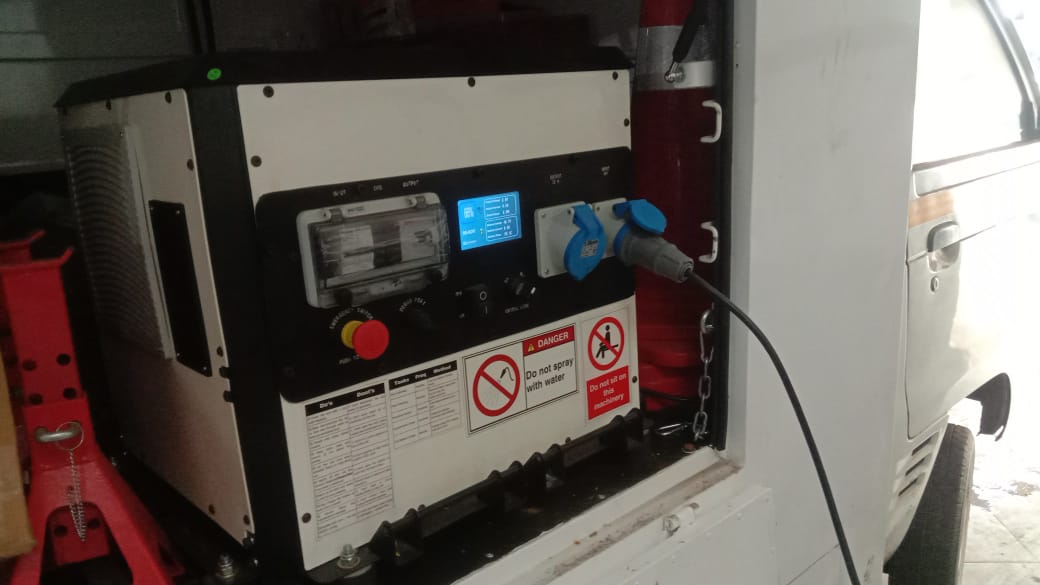 White industrial generator with control panel, red emergency stop button, and blue power plug connected inside a vehicle compartment.