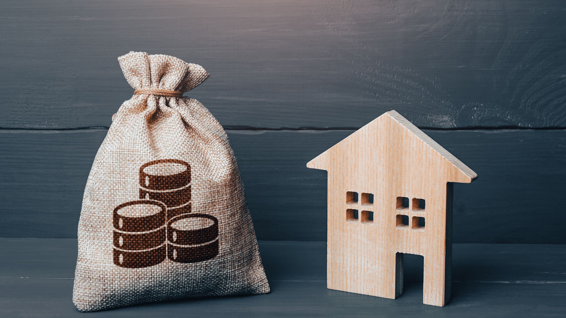 Stoffsack mit Münzsymbol neben einem kleinen Holzhaus als Symbol für Anlagevorschlag und Investition.