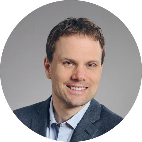 Porträt eines Mannes mit kurzen braunen Haaren und hellem Teint, der freundlich lächelt. Er trägt ein dunkles Sakko über einem hellen Hemd. Der Hintergrund ist neutral grau.