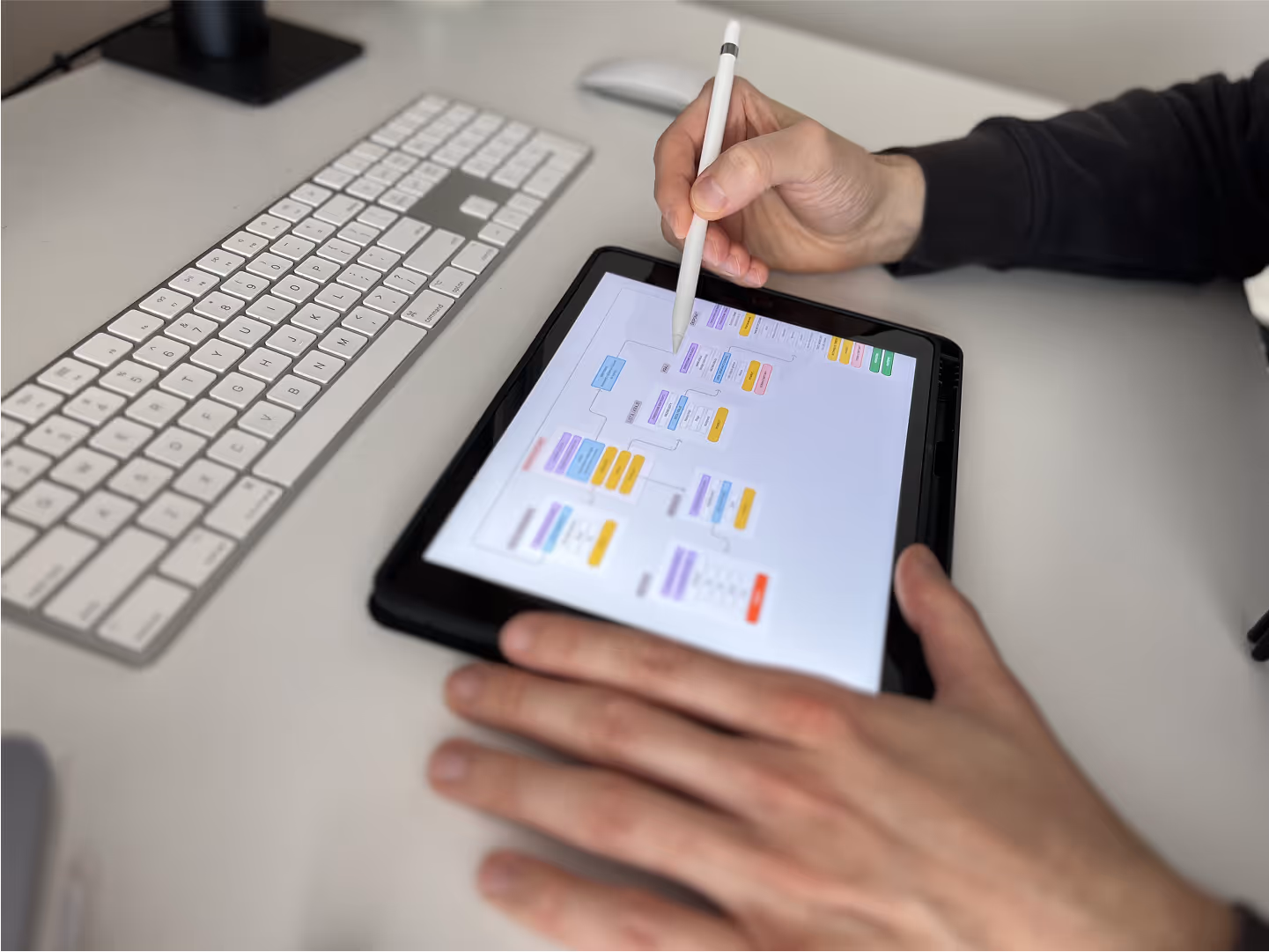 Dłonie Mateusza Mrozka pracującego nad projektem aplikacji. W prawej ręce trzyma Apple Pen, lewa na tablecie z wyświetloną architekturą informacji. Widoczna również klawiatura i myszka na biurku. Zdjęcie symbolizuje proces projektowania UX.