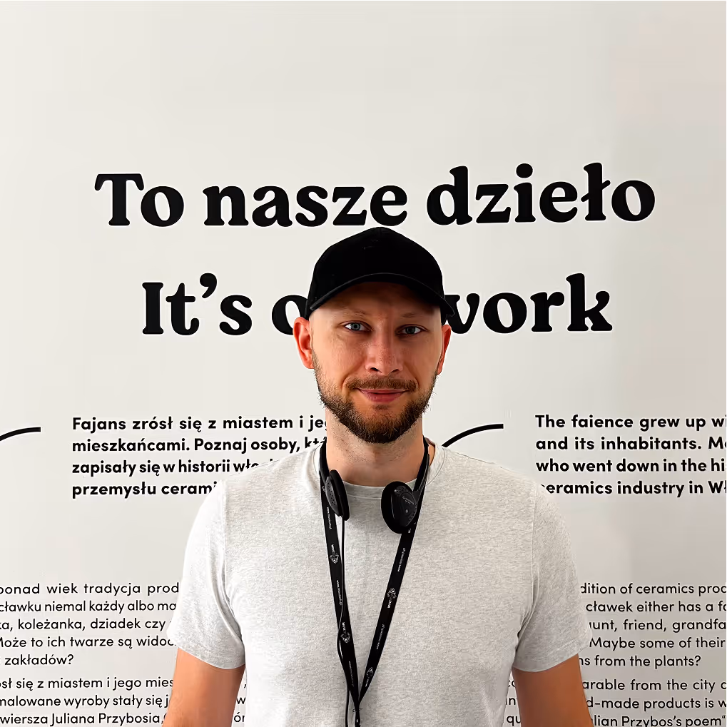 Mateusz Mrozek na tle ściany w Skarbcu Fajansu we Włocławku, prezentujący zaprojektowane przez siebie napisy i elementy graficzne wystawy muzealnej.