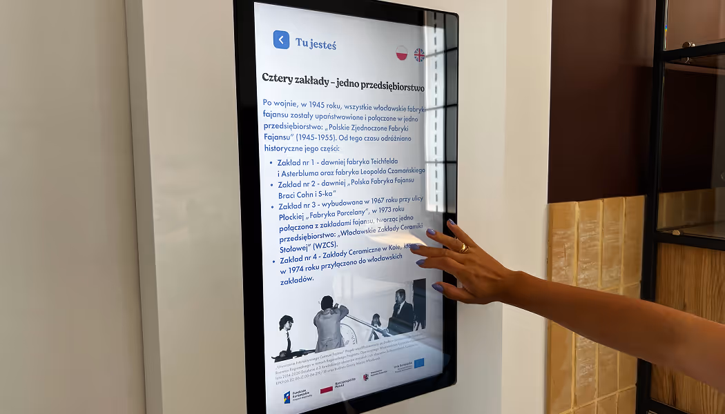 Ekran z aplikacją muzealną uzupełniającą wystawę w Skarbcu Fajansu. Widoczna dłoń zwiedzającej przesuwająca palec po ekranie dotykowym, demonstrująca interakcję UX.