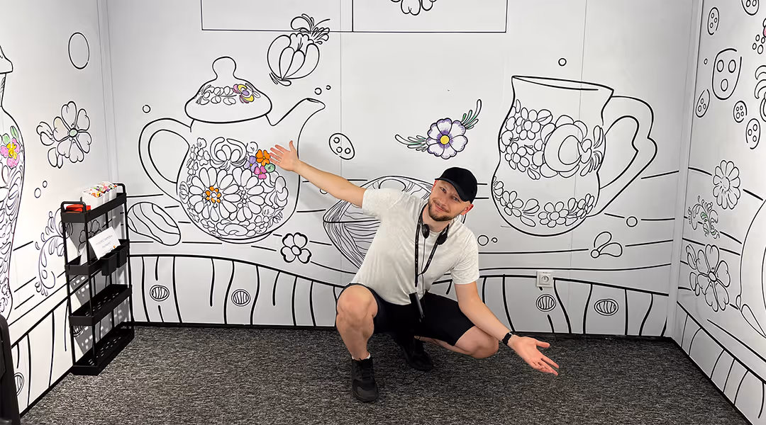 Mateusz Mrozek pozujący na tle zaprojektowanej przez siebie kolorowanki XXL na wystawie Skarbiec Fajansu. Zdjęcie ilustrujące autorstwo projektu.