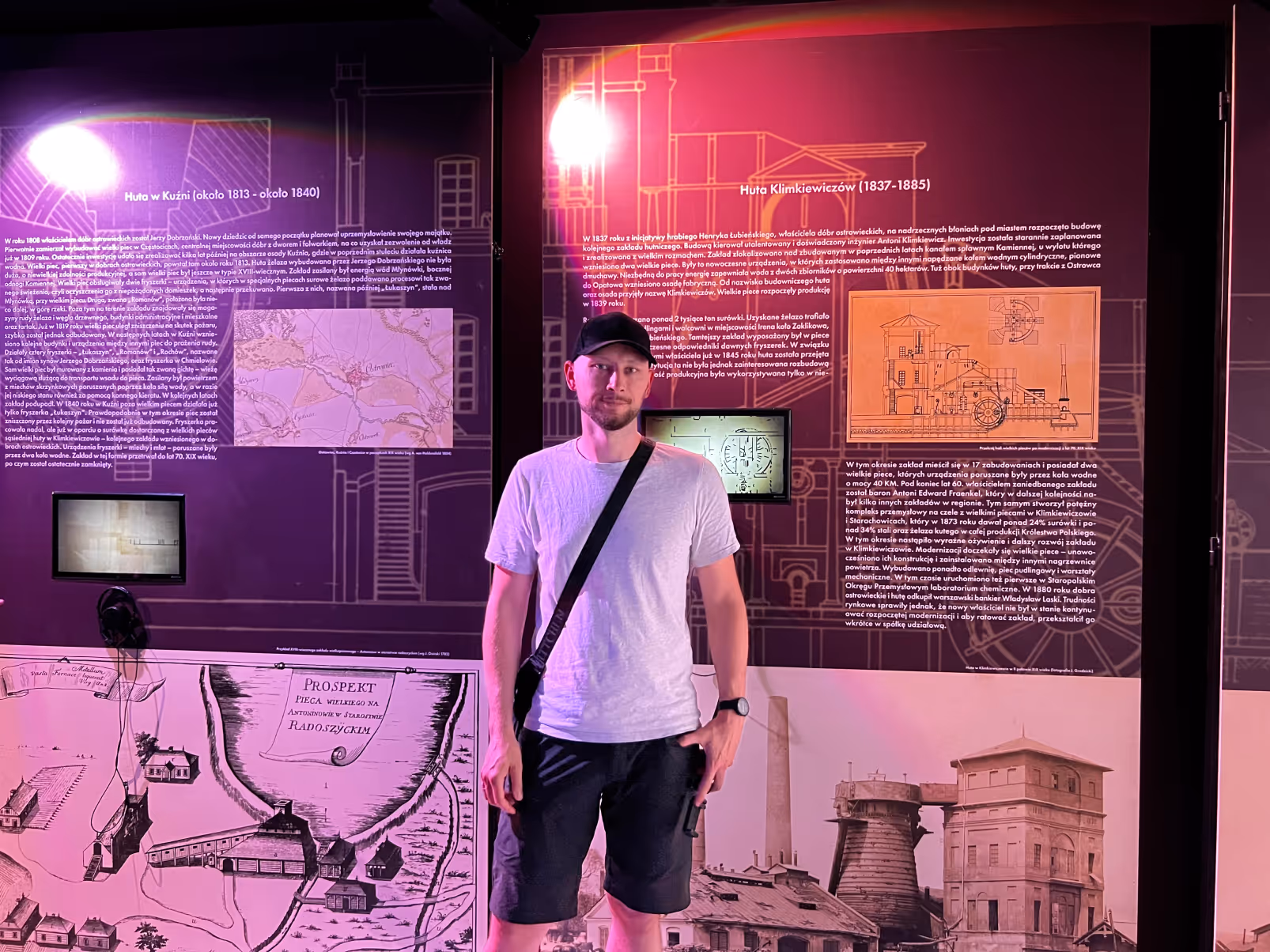 Mateusz Mrozek pozujący na tle wielkoformatowych infografik na ścianach Centrum Tradycji Hutnictwa. Zdjęcie ilustrujące autorstwo projektu graficznego wystawy muzealnej.