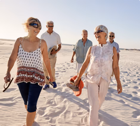 A group of four smiling senior adults enjoying a walk along a beach, with two women in the foreground leading the way and two men chatting in the background under a clear blue sky.