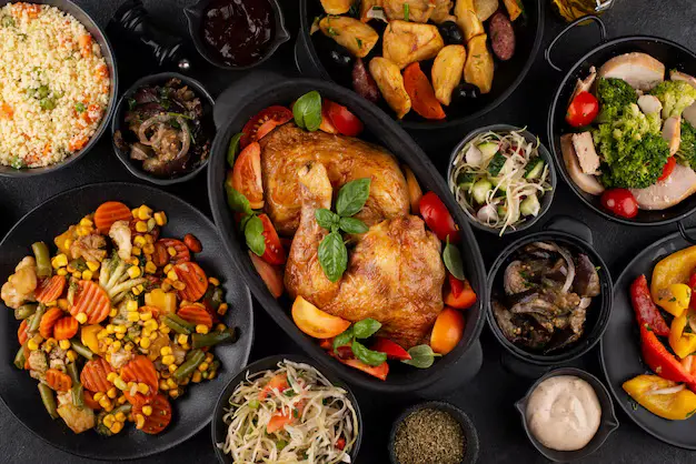 Top view of a table filled with a roasted whole chicken garnished with basil and cherry tomatoes, surrounded by various side dishes including grilled vegetables, salads, sauces, and couscous.