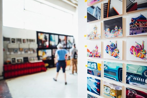 Art gallery with colorful photographs displayed on a white wall and two visitors observing artwork in the background.