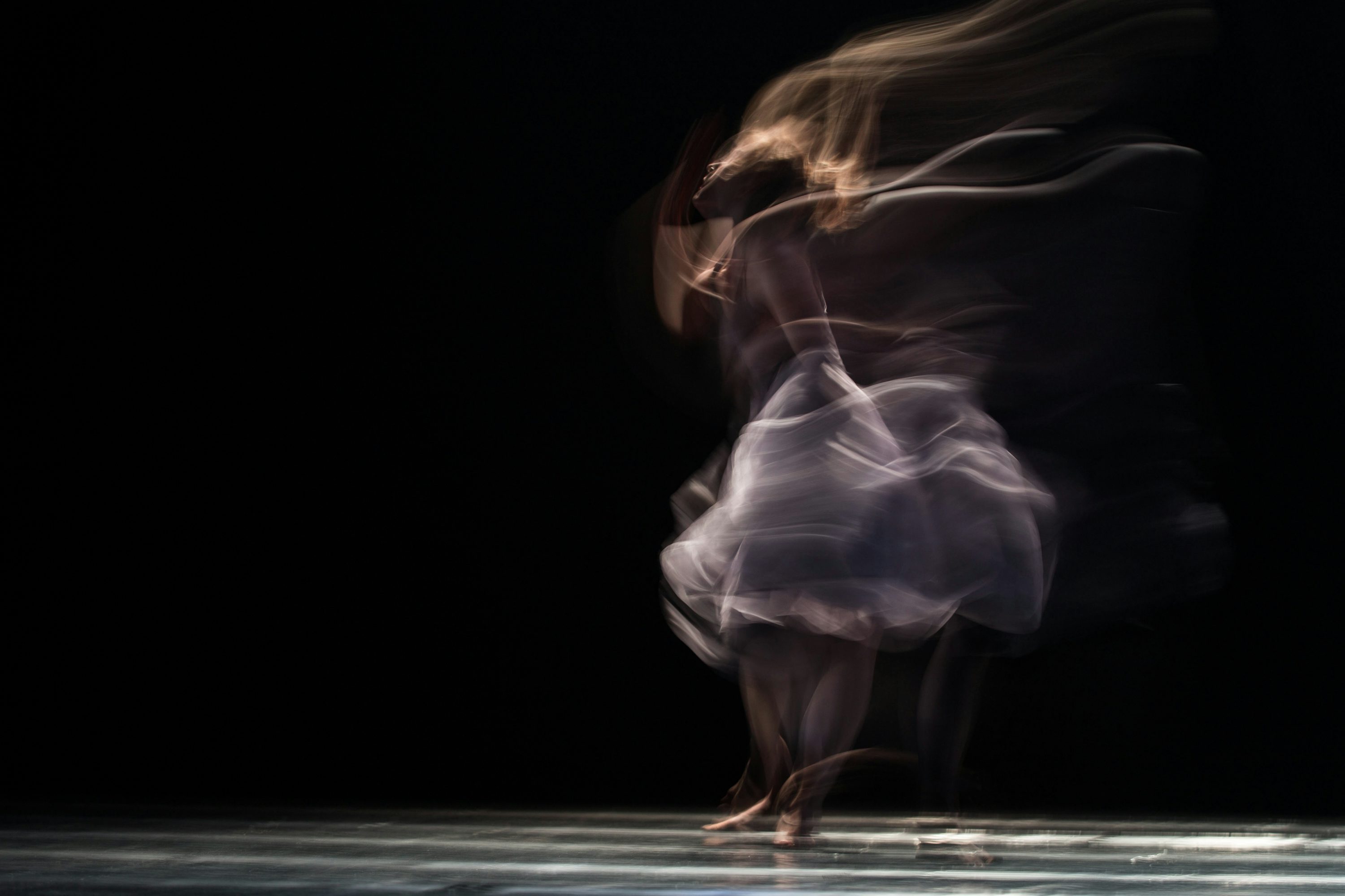 Blurred image of a ballet dancer spinning on stage with flowing dress and hair in motion against a black background.