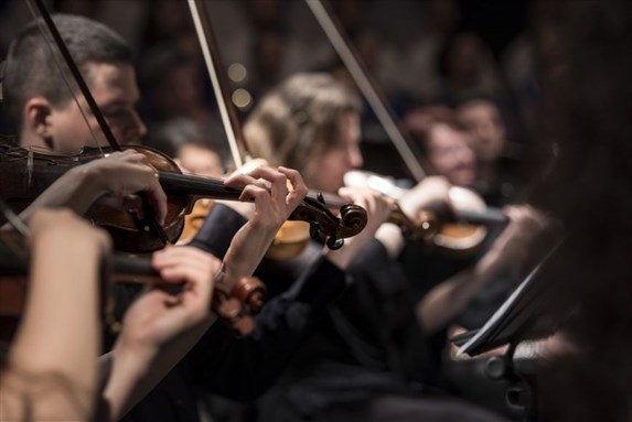 Musicians playing violins in an orchestra with a blurred background.