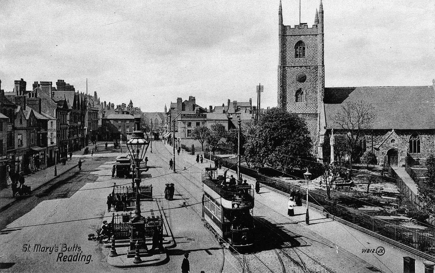 Black and white photo of a tram on tracks passing St. Mary's Butts church in Reading with people walking on the street and historic buildings lining the road.