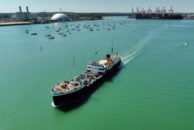 Large passenger ship sailing in a harbor with anchored sailboats and industrial buildings in the background.