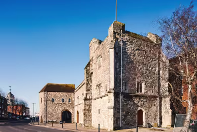 Historic stone castle building under clear blue sky with tree and surrounding pavement.