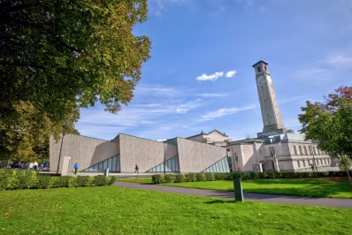 Modern angular building next to a tall clock tower surrounded by green lawns and trees under a blue sky.