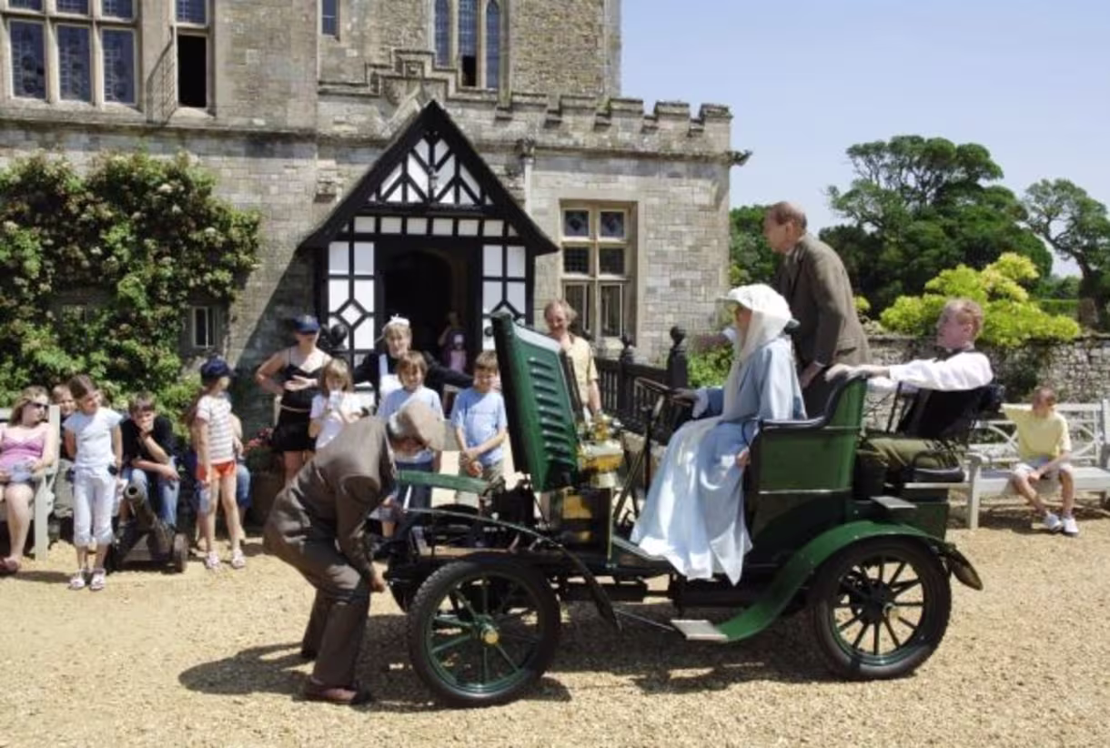 People dressed in historical clothing around an early vintage car in front of a stone building with onlookers watching.