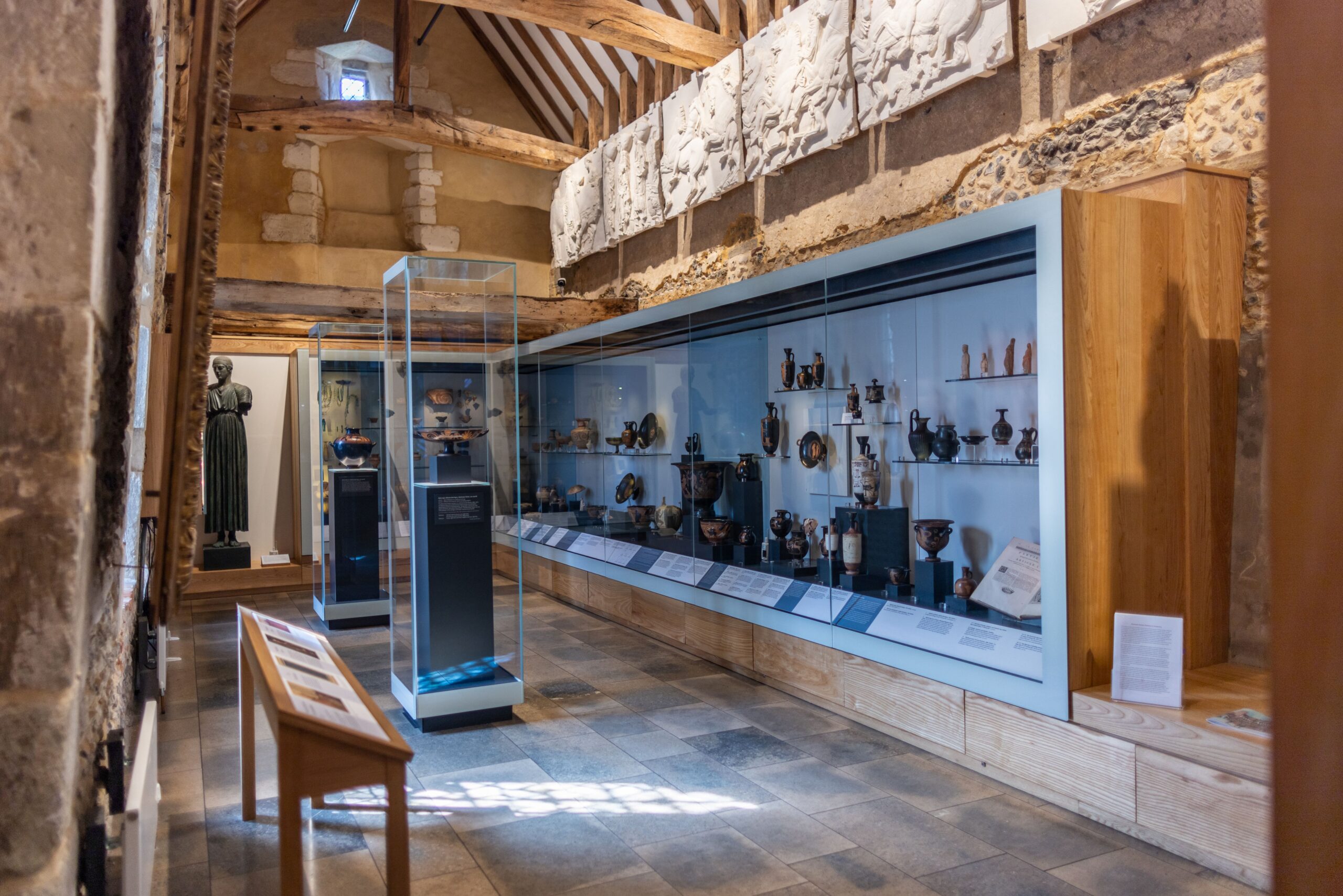 Museum gallery with ancient pottery and artifacts displayed in glass cases under wooden beams and stone walls.