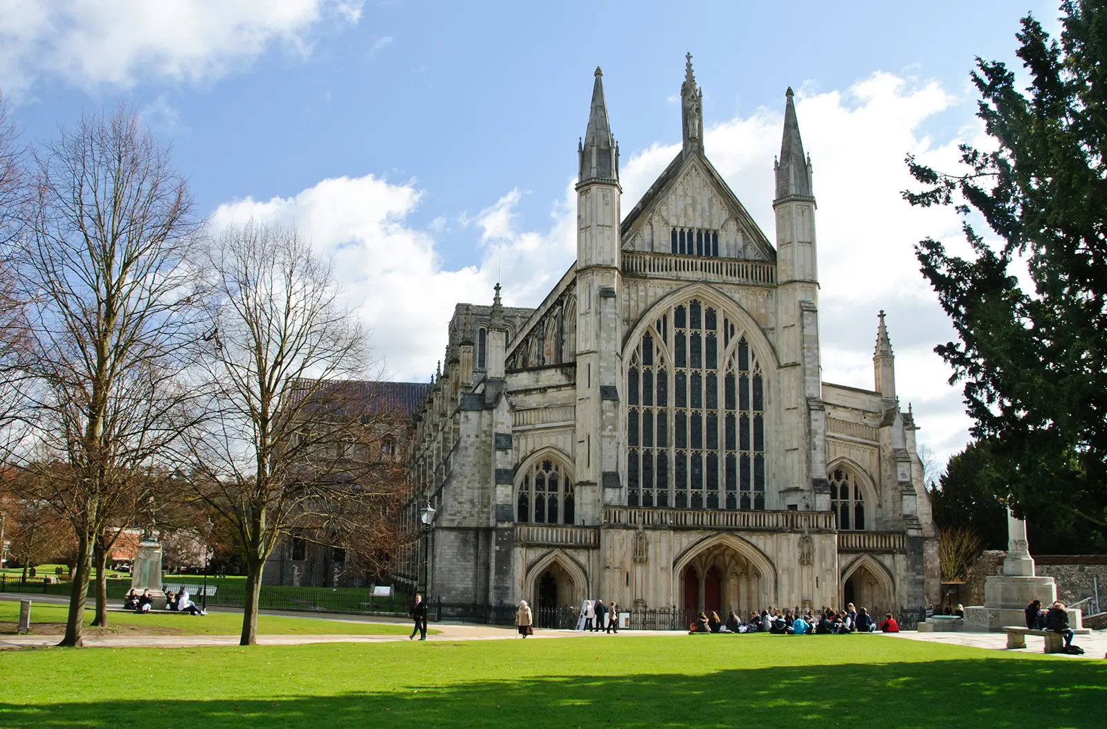 Winchester Cathedral with people sitting and walking on the surrounding green lawn under a partly cloudy blue sky.